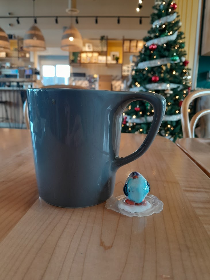 Custom penguin figurine, hand-sculpted and painted, displayed on a coffee shop background. Finished clay figurines created by students in Goldfish Art’s clay sculpting and character modeling course for ages 5-18